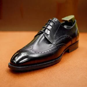 May include: A single black leather dress shoe with a classic wingtip design. The shoe features intricate perforations along the seams and toe, and is laced up with black laces. The shoe is set against a warm, neutral background.