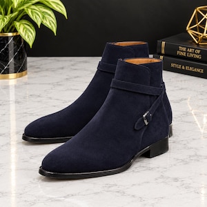 Könnte beinhalten: Ein Paar marineblaue Wildleder-Stiefeletten mit abgerundeter Zehenpartie, niedrigem Blockabsatz und einem dekorativen Riemen, der um den Knöchel verläuft und mit einer kleinen silberfarbenen Metallschnalle an der Außenseite befestigt ist.