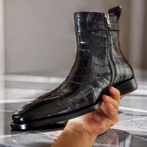May include: A black ankle boot with a glossy finish and a textured crocodile skin pattern. The boot features a pointed toe, a low block heel, and a silver-toned metal zipper running vertically along the side for closure and a pull tab at the top.