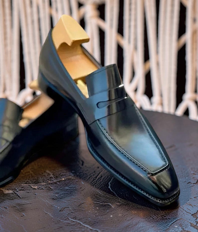 May include: Black leather penny loafers with a glossy finish. The shoes have a classic design with a square toe and a strap across the vamp. A wooden shoe tree is inserted inside the shoe. The loafers are on a dark surface.