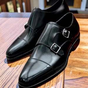 K&ouml;nnte beinhalten: Ein Paar schwarze Monk-Strap-Schuhe aus Leder mit silbernen Schnallen. Die Schuhe haben eine polierte Oberfl&auml;che und werden auf einer Holzoberfl&auml;che pr&auml;sentiert. Das Design verf&uuml;gt &uuml;ber eine Kappe und einen doppelten Schnallenverschluss.