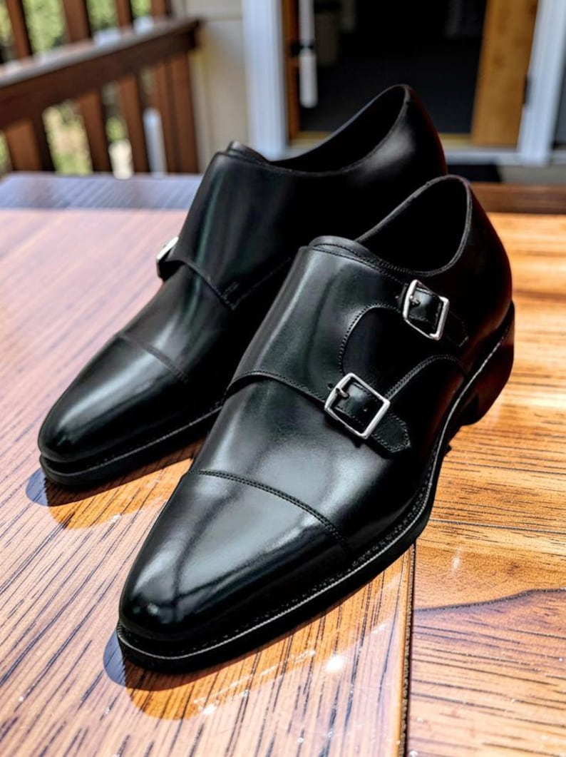 K&ouml;nnte beinhalten: Ein Paar schwarze Monk-Strap-Schuhe aus Leder mit silbernen Schnallen. Die Schuhe haben eine polierte Oberfl&auml;che und werden auf einer Holzoberfl&auml;che pr&auml;sentiert. Das Design verf&uuml;gt &uuml;ber eine Kappe und einen doppelten Schnallenverschluss.