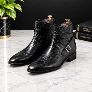 Puede incluir: Un par de botines negros con un patrón de piel de cocodrilo texturizado, una correa lateral con una hebilla de metal plateado y una horma de madera insertada en cada bota. Los botines tienen un acabado liso y pulido con un tacón bajo.