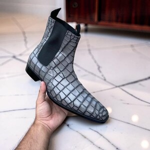 Peut inclure: Une bottine grise avec un motif texturé en peau de crocodile, des panneaux latéraux élastiques noirs et un talon noir. La botte présente un bout pointu, une silhouette élégante et des tirettes en haut de la tige pour faciliter l'enfilage.