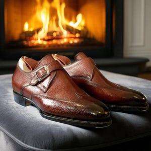 以下が含まれることがあります： バックル留めの茶色の革製ドレスシューズのペア。暖かく燃える火のある暖炉を背景にしています。靴は灰色の表面に置かれています。