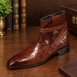Könnte beinhalten: Ein brauner Stiefeletten-Schuh mit glänzender Krokodilprägung im Leder. Der Schuh verfügt über einen dekorativen Riemen am Knöchel, der mit einer kleinen Metallschnalle befestigt ist, eine spitz zulaufende Zehenpartie und einen flachen schwarzen Absatz.