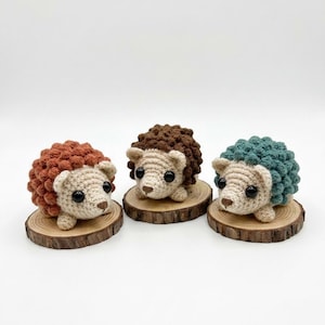 May include: Three crocheted hedgehogs in rust, brown, and teal, each with a beige face and black eyes. They sit on small, round wooden slices. The hedgehogs have textured, spiky backs, and are displayed against a white background.