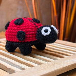 May include: A handmade crochet ladybug toy. The ladybug has a red body with black spots, a black head, and black legs. The eyes are white with black pupils. The toy is sitting on a wooden slatted surface.