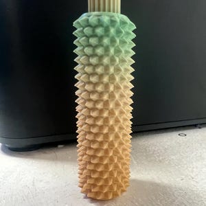Puede incluir: Un tapón de botella decorativo con un diseño geométrico texturizado. El tapón presenta un degradado de colores, que va del verde al marrón claro. La parte superior tiene un diseño acanalado.