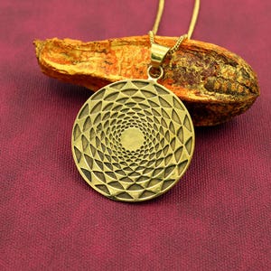 Pode incluir: Um colar com pingente dourado com um design circular com um padrão em espiral. O pingente é suspenso por uma corrente delicada. O colar repousa sobre uma superfície roxo-avermelhada, com um pedaço de casca marrom-alaranjada seca ao fundo.