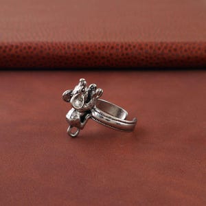 May include: A silver-colored ring featuring a detailed mouse design. The mouse is sculpted in a playful pose, with its arms raised. The ring has an adjustable band for a comfortable fit. The ring is set against a brown background.