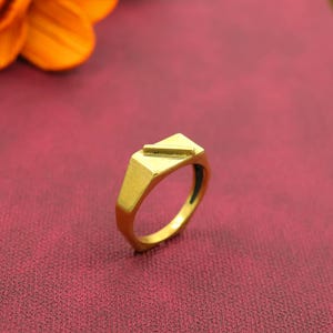 Könnte beinhalten: Ein goldfarbener Ring mit geometrischem Design. Der Ring hat eine dreieckige Oberseite mit einem horizontalen Balken. Das Band hat ein facettiertes Design. Der Ring ist vor rotem Hintergrund platziert.