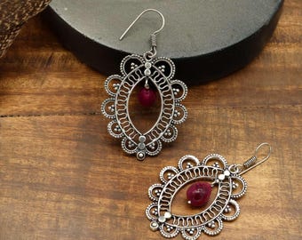 May include: Two silver-toned earrings with an oval filigree design and a small, red gemstone in the center. The earrings have a floral-like border and a hook closure. They are displayed on a wooden surface.