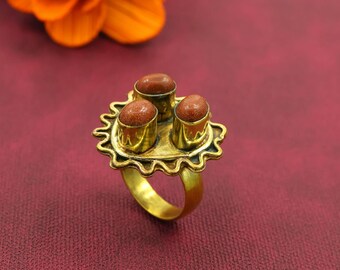 May include: A gold ring with three brown gemstones set on a decorative, sunburst-shaped base. The ring has a thick band and a detailed, textured design. The background is a deep red with an orange flower in the background.