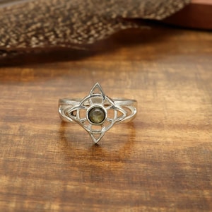 Puede incluir: Un anillo de plata con un diseño intrincado y calado. El anillo presenta un engaste central circular con una pequeña piedra redonda de color verde oliva. El diseño incluye una forma de estrella de cuatro puntas y patrones geométricos. El anillo se muestra sobre una superficie de madera.