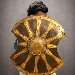 May include: A circular shield with a scalloped edge and two side cutouts, featuring a central gold-toned medallion with intricate floral engravings. The surface displays alternating dark brown and raised gold-toned radial panels with metal rivets.