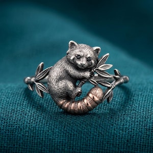 Op de afbeelding: Een zilverkleurige ring met een sculptuur van een rode panda die in een bamboetak klimt. De panda heeft donkere ogen en een gestreepte staart met een koperkleurige afwerking. De ringband is vormgegeven als bamboestengels.