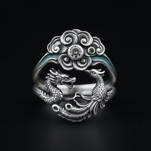 May include: A silver ring featuring a sculpted dragon and phoenix facing each other. The top section displays a cloud motif with a central clear round gemstone and a small green accent stone. The band includes teal enamel detailing along the sides.