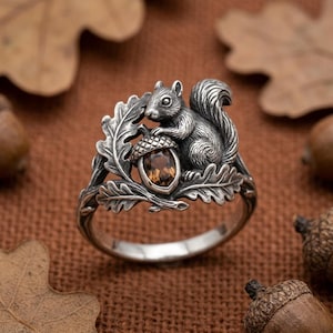 May include: A silver ring featuring a detailed squirrel perched on an oak leaf branch. The squirrel holds a small acorn with a faceted, dark brown gemstone set into the cap. The metal has an oxidized finish to highlight the textured details.