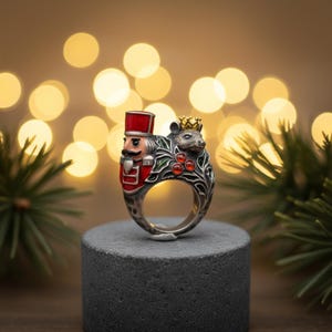 Peut inclure: Bague en argent représentant un soldat Casse-Noisette et une souris couronnée. Le Casse-Noisette porte un chapeau et un manteau rouges, tandis que la souris porte une couronne dorée. La bague est ornée de gemmes rouges et de feuilles vertes.