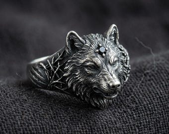 May include: A silver-toned ring featuring a detailed wolf head sculpture with a small, round, black gemstone set between the ears. The band of the ring displays an intricate, raised relief pattern of bare tree branches extending from the wolf head.