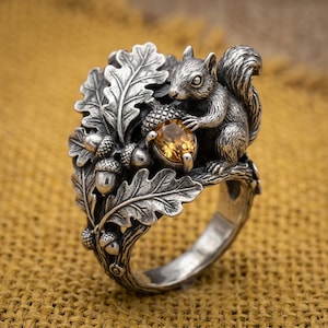 May include: A silver ring featuring a detailed squirrel perched on an oak branch with textured leaves and several small acorns. An oval-shaped, golden-yellow gemstone is set within an acorn cap held by the squirrel, showcasing intricate metalwork.