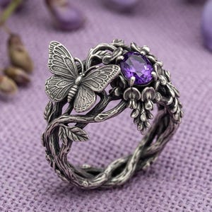 Puede incluir: Un anillo de plata con una mariposa detallada posada sobre una banda con textura de rama. El diseño incluye pequeñas hojas y una piedra preciosa ovalada de color púrpura engastada entre elementos florales esculpidos.