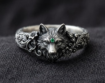 May include: A silver ring featuring a detailed fox head at the centre, accented with a small green gemstone set between the eyes. The band is decorated with intricate floral and vine engravings that extend from the fox head along the sides.