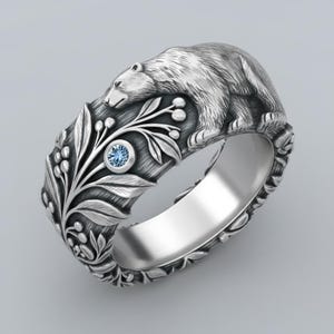 May include: Silver ring featuring a detailed bear design. The ring has a floral pattern with a small, round, light blue gemstone. The bear is sculpted on the top of the ring, with the floral design wrapping around the band.