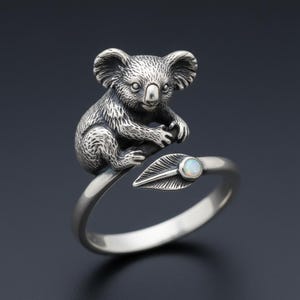 May include: A silver-toned ring featuring a detailed koala bear perched on a leaf. The ring has an open design, with a small, round, opalescent stone set in the leaf. The koala's fur and features are intricately rendered.