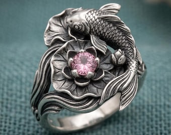 May include: A silver ring featuring a sculpted koi fish wrapping around a central lotus flower. The lotus flower holds a round, faceted pink gemstone at its centre. The metal has an oxidised finish to highlight the detailed scales and petals.