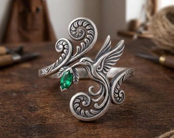 May include: A silver-toned metal cuff bracelet featuring an intricate hummingbird design with engraved scrollwork patterns. The bird is depicted in flight, holding a single marquise-cut emerald green gemstone in its beak, creating a detailed focal point.