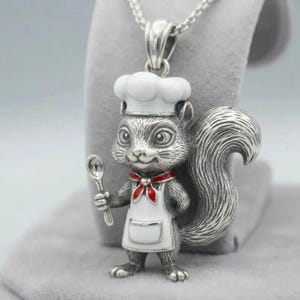May include: A silver-colored pendant necklace featuring a squirrel chef. The squirrel wears a white chef's hat, apron, and a red bow tie, holding a spoon. The pendant is attached to a silver chain.