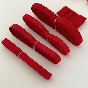 May include: A selection of red bra-making supplies. The image features several spools of red elastic, some with a decorative scalloped edge, and a red bra hook and eye closure. The items are arranged on a white surface.