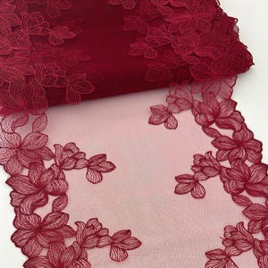 Op de afbeelding: Bordeaux bloemen kanten stof. De stof heeft een gedetailleerd bloemenontwerp met een mesh rug. De bloemen zijn geborduurd in een dieprode tint, waardoor een delicate en elegante uitstraling ontstaat. De stof is gedeeltelijk opgerold, wat de textuur en diepte laat zien.