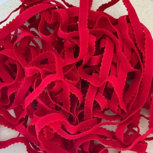 May include: A close-up of a pile of bright red, scalloped-edge elastic ribbon. The ribbon is soft and flexible, with a slightly textured surface. The edges have a decorative, wavy pattern. Suitable for sewing and crafting projects.