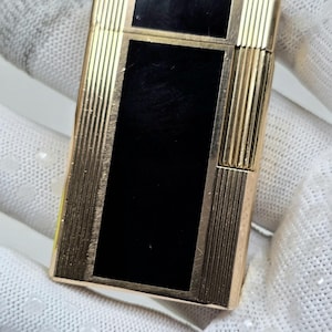May include: A gold-coloured lighter featuring a central rectangular black panel. The sides of the lighter have vertical ribbed detailing and a polished finish. The lighter is held in a white gloved hand.