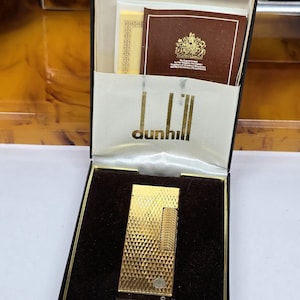 May include: A gold-tone metal lighter with a textured grid pattern, presented in a black hinged box. The interior lid features the white text dunhill and a brown card with a gold royal crest. The lighter rests on a dark brown velvet base.