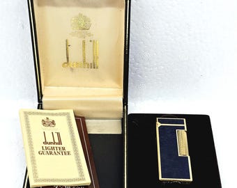 May include: A gold and dark blue rectangular lighter with a textured finish, presented in a black hinged box with a cream interior. The set includes a cream-colored paper booklet labeled Dunhill Lighter Guarantee and a dark brown card.