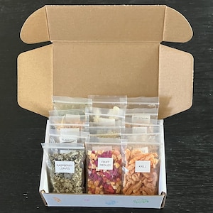 May include: An open cardboard box containing small, clear plastic bags filled with various dried ingredients. The bags are labeled with text such as "Raspberry Leaves," "Fruit Medley," and "Krill." The box sits on a dark surface.