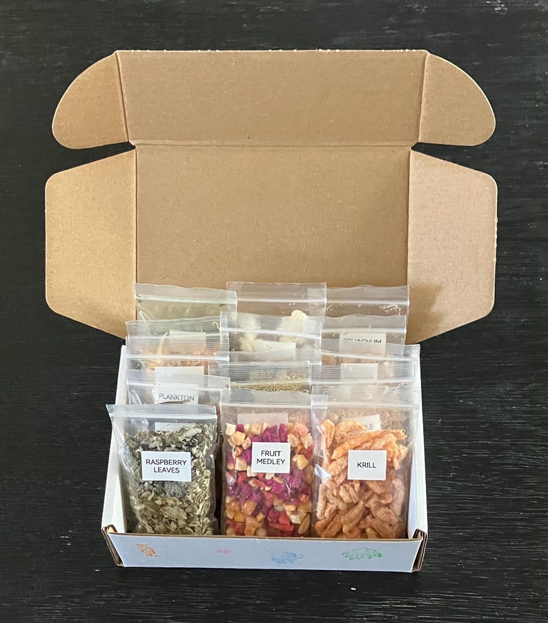 May include: An open cardboard box containing small, clear plastic bags filled with various dried ingredients. The bags are labeled with text such as "Raspberry Leaves," "Fruit Medley," and "Krill." The box sits on a dark surface.