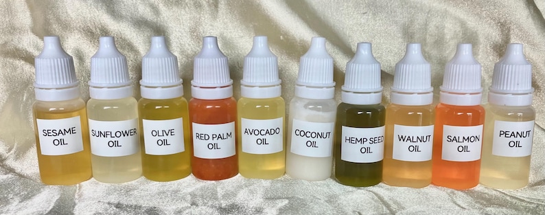 May include: A row of ten small clear bottles with white caps and labels. Each bottle contains a different colored oil, including sesame, sunflower, olive, red palm, avocado, coconut, hemp seed, walnut, salmon, and peanut oil.