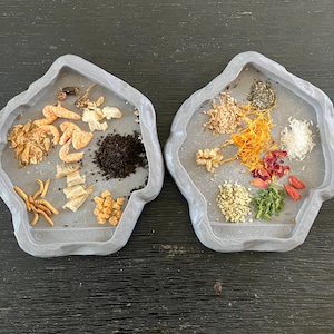 May include: Two gray, irregularly shaped trays filled with various dried ingredients. One tray contains dried shrimp, insects, and dark granules. The other tray holds seeds, herbs, and dried flowers. The trays are set against a dark wood-grain surface.
