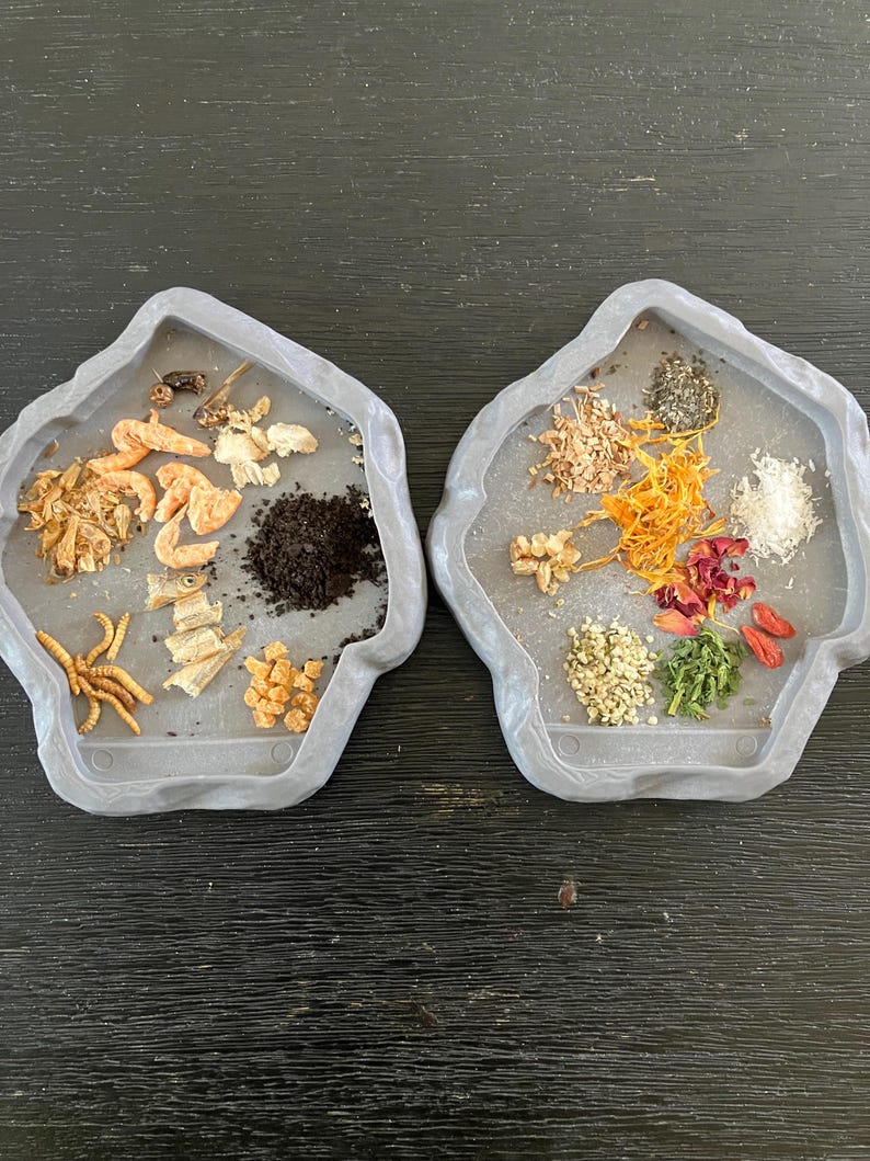 May include: Two gray, irregularly shaped trays filled with various dried ingredients. One tray contains dried shrimp, insects, and dark granules. The other tray holds seeds, herbs, and dried flowers. The trays are set against a dark wood-grain surface.