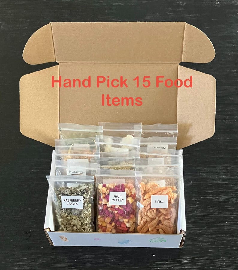 May include: An open cardboard box filled with 15 small, clear plastic bags of food items. The bags are labeled with names like "Raspberry Leaves," "Fruit Medley," and "Krill." The box has the text "Hand Pick 15 Food Items" in red.