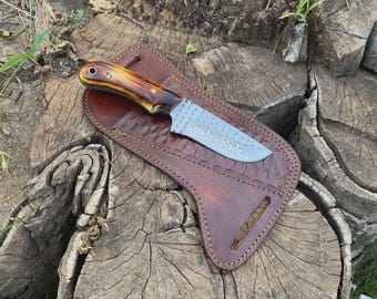 May include: A handcrafted knife with a wooden handle and a silver blade, in a brown leather sheath. The blade is engraved with the word "Cowboyblade". The sheath has decorative stitching.