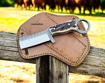 Peut inclure: Un couteau artisanal avec une lame en acier inoxydable et un manche en bois de cerf. Il repose sur un étui en cuir marron avec des coutures décoratives. Le texte "CowboyBlade" est visible sur l'étui.