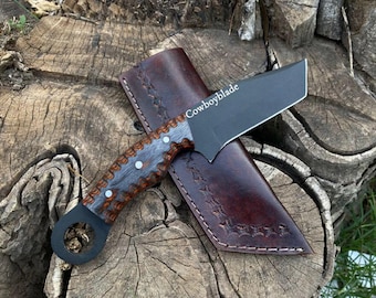May include: A black knife with the text "Cowboyblade" on the blade, resting on a brown leather sheath. The handle is gray and brown with white accents. The knife and sheath are on a wooden surface.