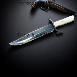 May include: A large, polished steel knife with a cream-colored handle and a curved guard. The blade is sharp and reflective. A vintage firearm is in the background. The text "Cowboy Blade" is visible.