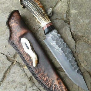 Puede incluir: Un cuchillo artesanal con una hoja texturizada y un mango de asta. El cuchillo se presenta con una funda de cuero marrón. La hoja tiene el texto "Bales Casas".